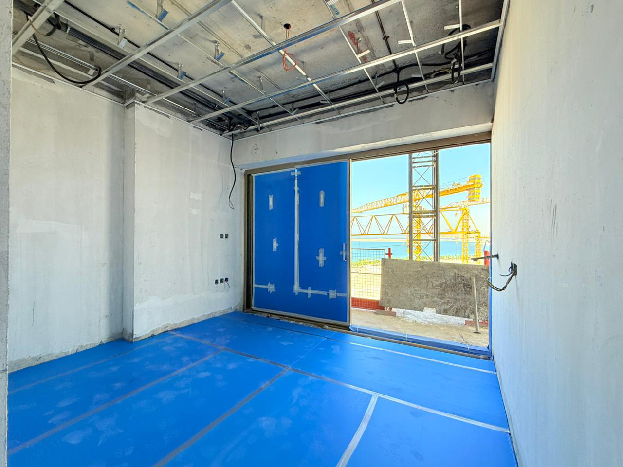Beach Walk Iii By Imtiaz An under-construction room with concrete walls, blue flooring, and a large window showing industrial cranes outside.